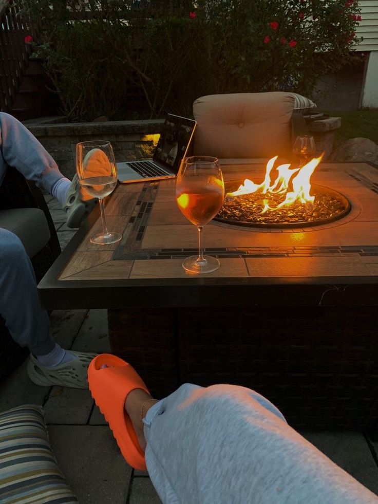 two people sitting at a fire pit with wine glasses on the table next to them