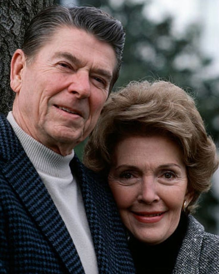 an older man and woman standing next to each other near a tree with their arms around each other