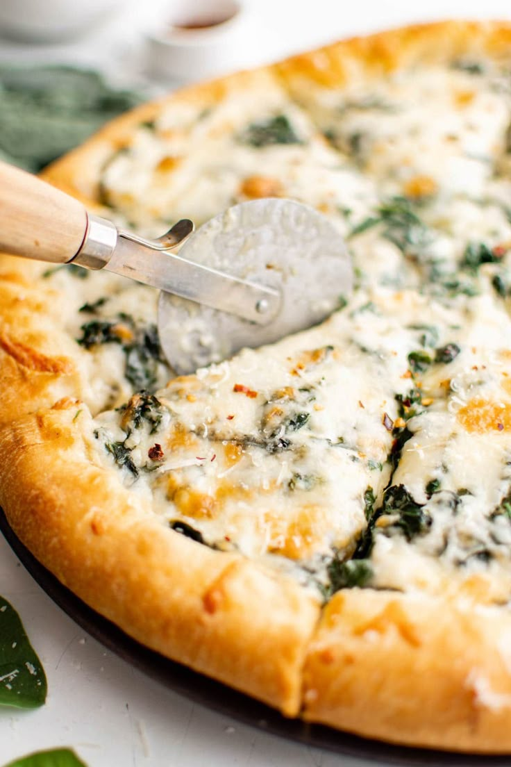 a pizza with cheese and spinach on it being cut by a pizza peeler