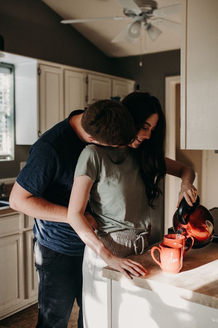 a man and woman standing in a kitchen next to an orange coffee mug on a counter