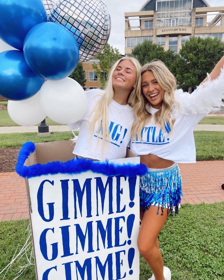 two women in cheerleader outfits standing next to a giant gummie sign with blue and white balloons