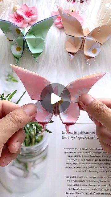 two hands holding origami butterflies in front of an open book with pink flowers