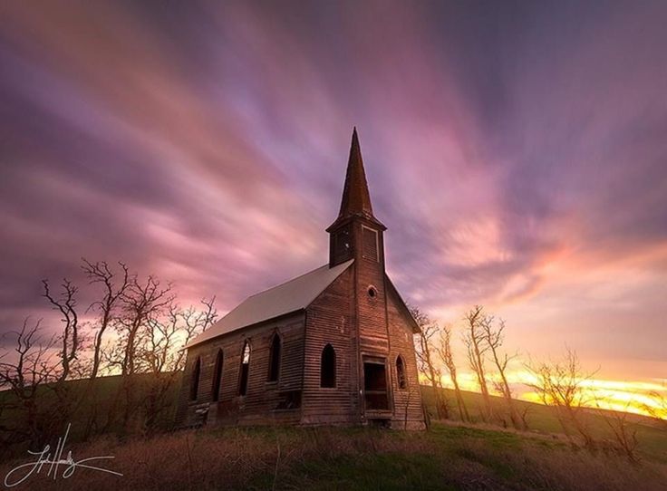 an old church sits in the middle of a grassy field with trees around it at sunset