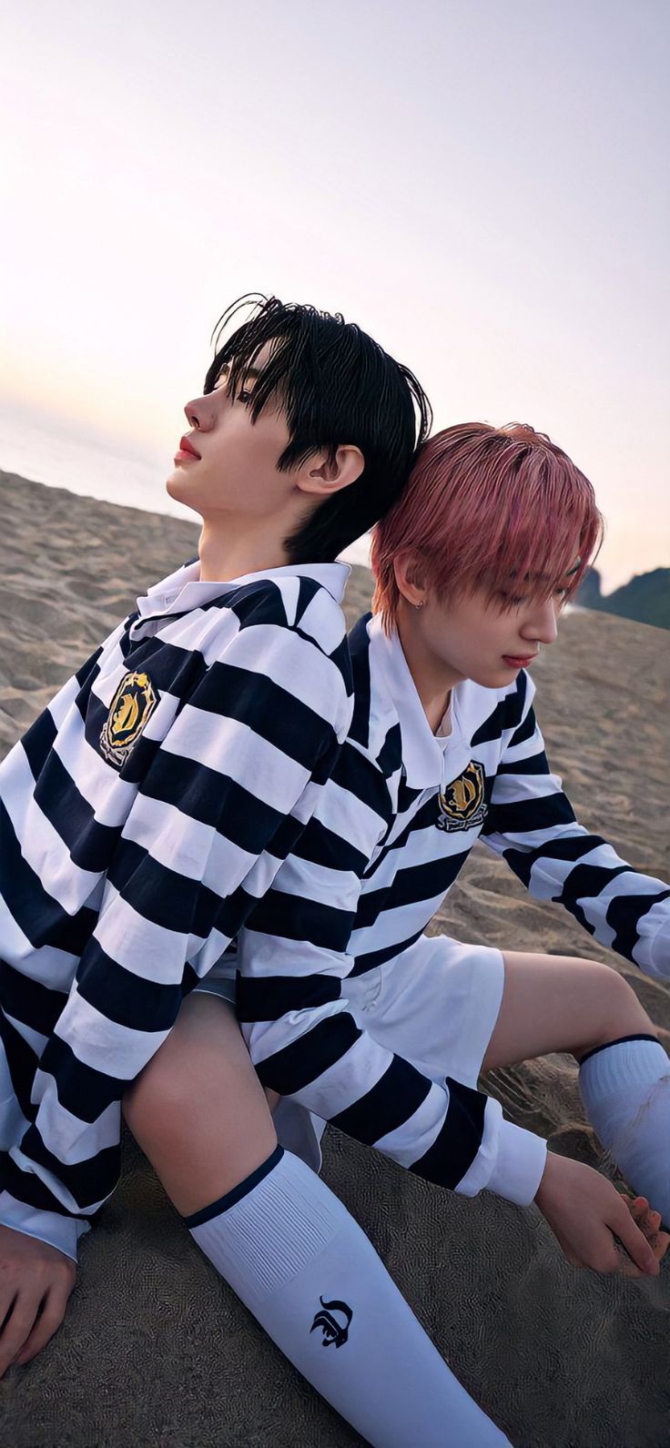 two young men sitting on the sand at the beach, one wearing a striped shirt