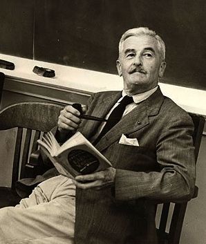 black and white photograph of man in suit sitting on chair with book open, looking at camera