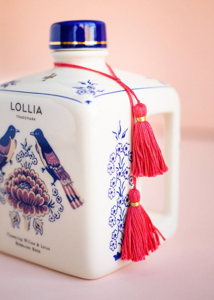 a small white box with red tassels and birds painted on the side, sitting on a pink surface