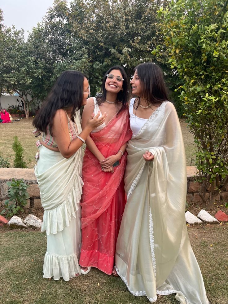 three women in sari standing next to each other on the grass with trees behind them