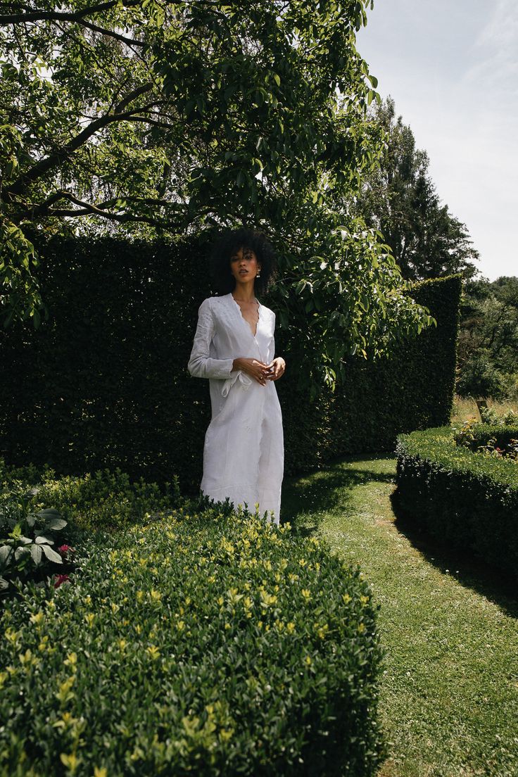 a woman standing in the middle of a lush green field next to bushes and trees