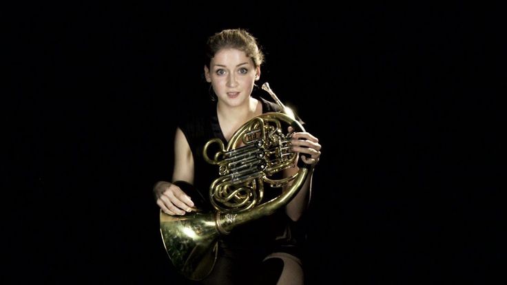 a woman holding a french horn in her right hand and posing for the camera on a black background