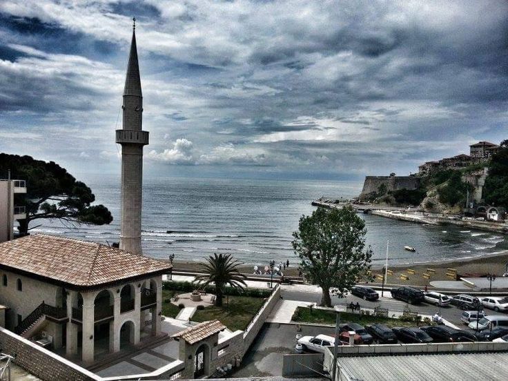 an old building with a steeple next to the ocean and cars parked in front of it