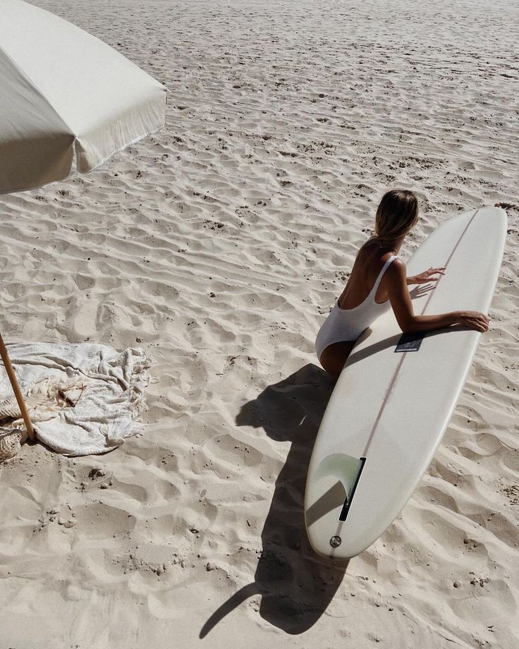 a woman is sitting on the beach with her surfboard under an umbrella and looking at the ocean