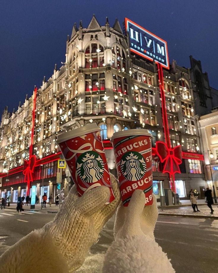 someone holding up two cups of starbucks coffee in front of a building with lights on it