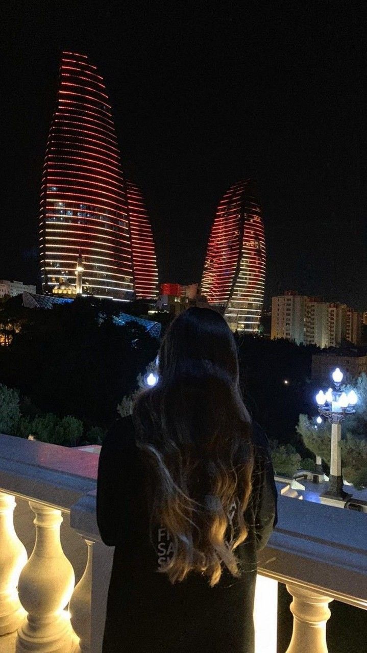 a woman standing on top of a balcony next to a railing at night with the city lights in the background