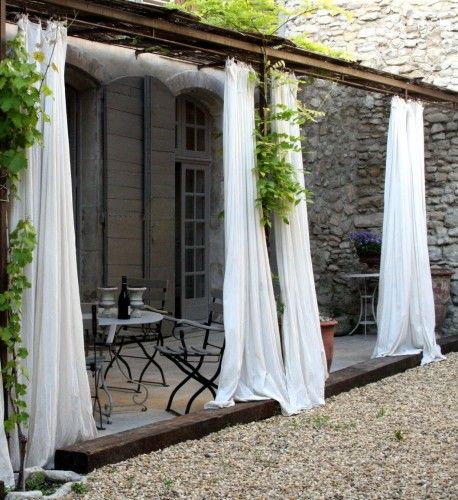 an outdoor patio covered in white drapes and plants next to a table with chairs