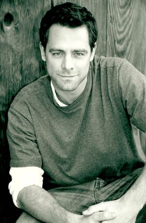 a black and white photo of a man sitting in front of a wooden wall with his hands on his knees