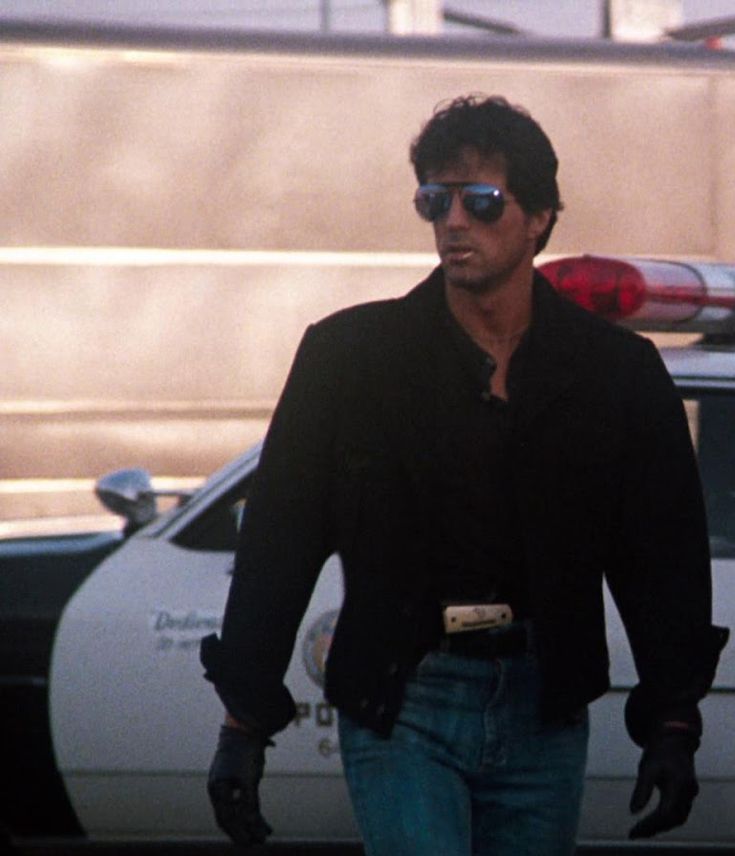 a man walking down the street in front of a police car with sunglasses on his head