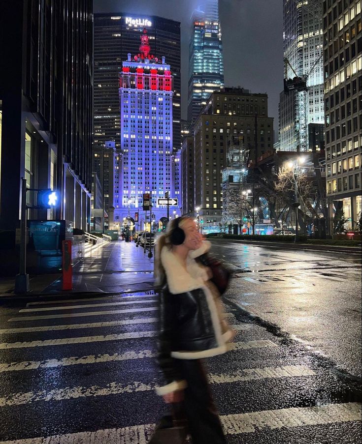 a woman walking across a cross walk in front of tall buildings with lights on at night