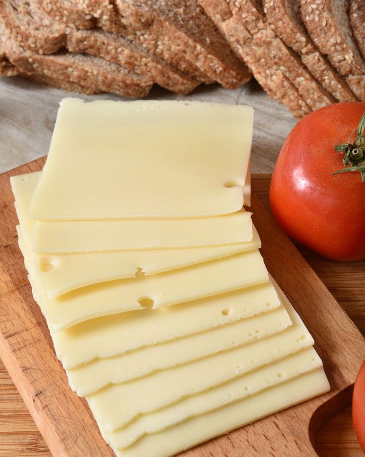 several slices of cheese on a cutting board with tomatoes and bread in the back ground