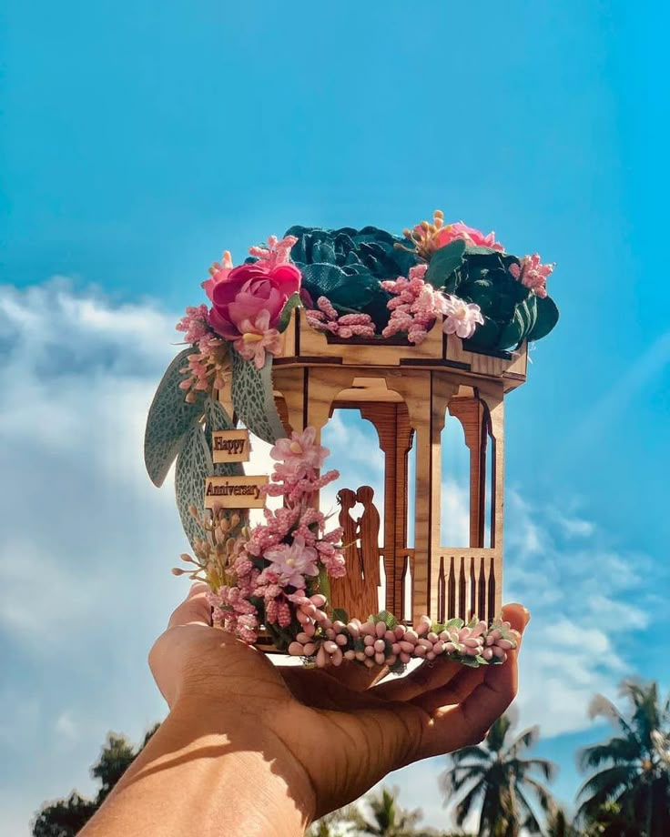 a hand holding up a small wooden birdhouse with flowers on the roof and palm trees in the background