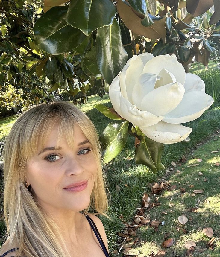 a beautiful blonde woman standing next to a white flower in the grass and looking at the camera