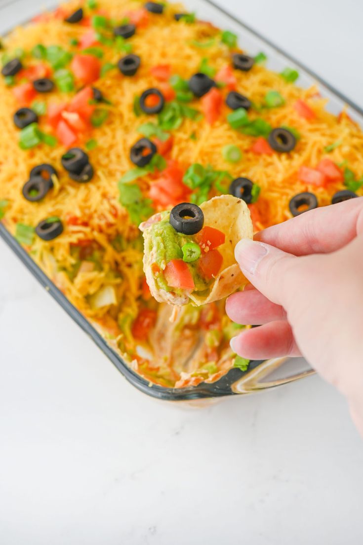 a hand holding a tortilla chip with black olives, cheese and guacamole