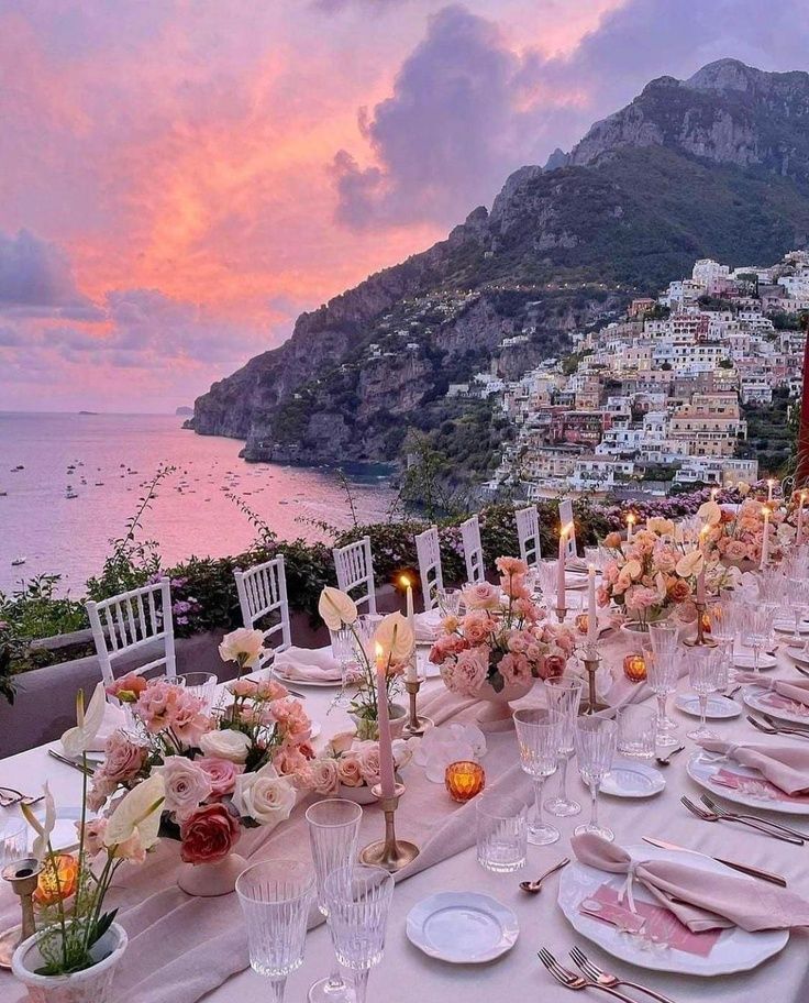 a table set up with flowers and candles for an outdoor dinner overlooking the ocean at sunset