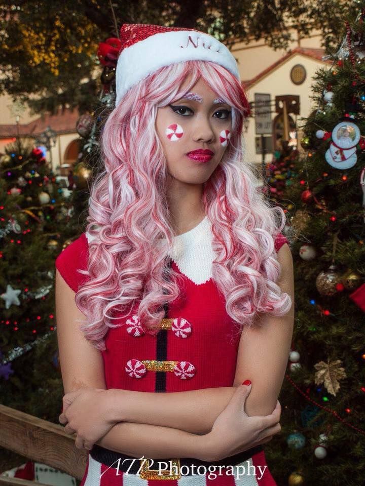 a woman with long pink hair wearing a santa hat and red dress standing in front of a christmas tree