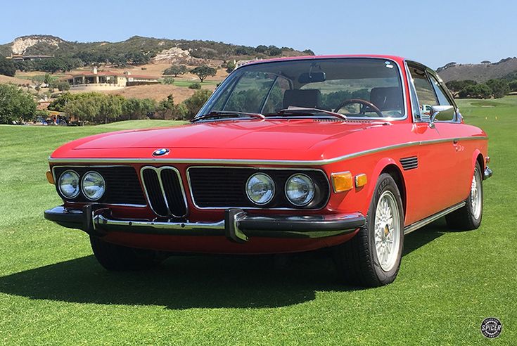 an old red bmw is parked on the grass in front of some hills and trees