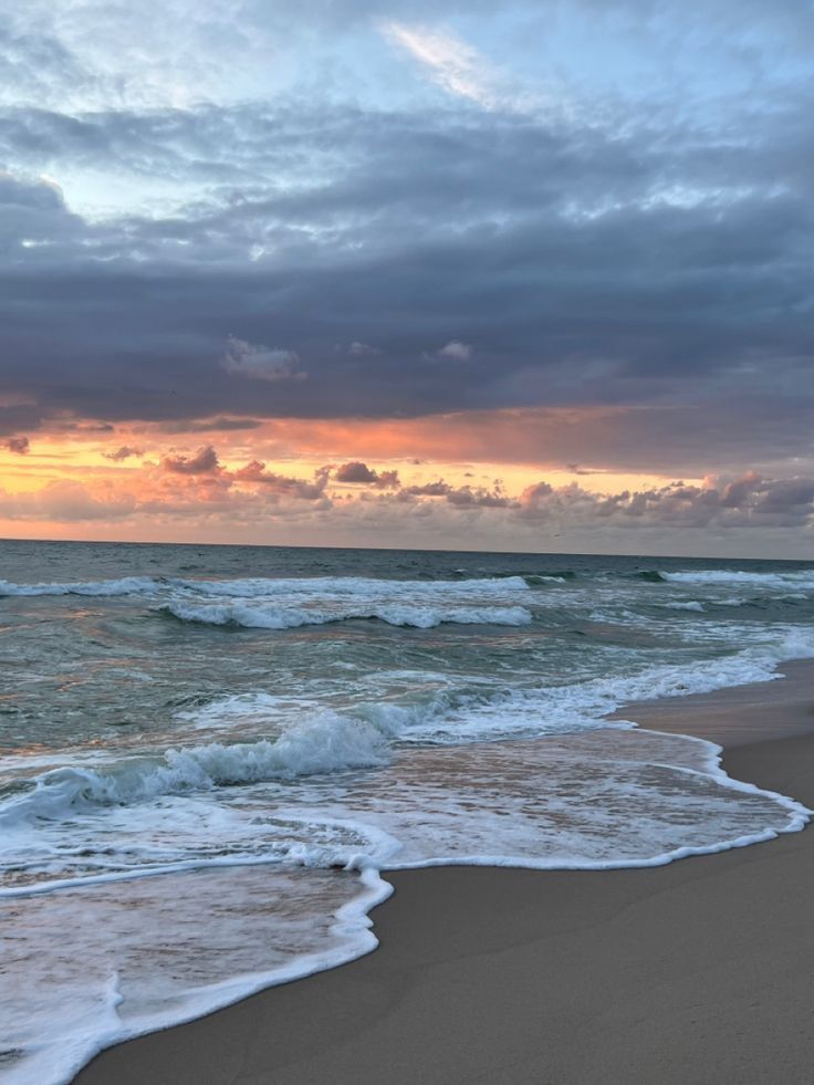 the sun is setting over the ocean with waves crashing on the beach and clouds in the sky