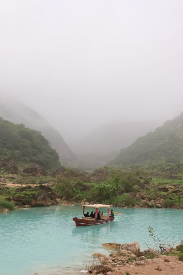 a small boat floating on top of a lake surrounded by green mountains in the fog