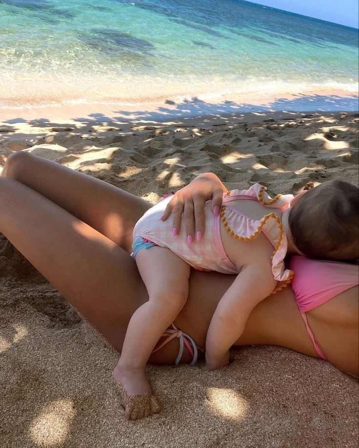 a woman laying on top of a sandy beach next to the ocean holding a baby