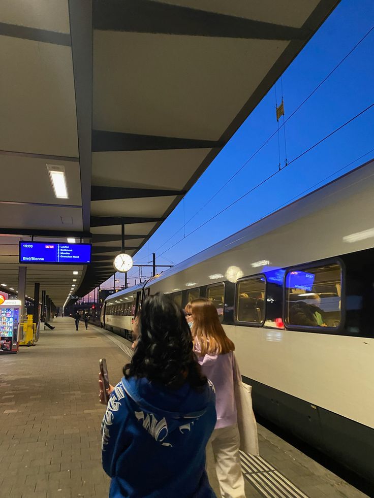 two women standing next to each other near a train at a station with blue sky in the background