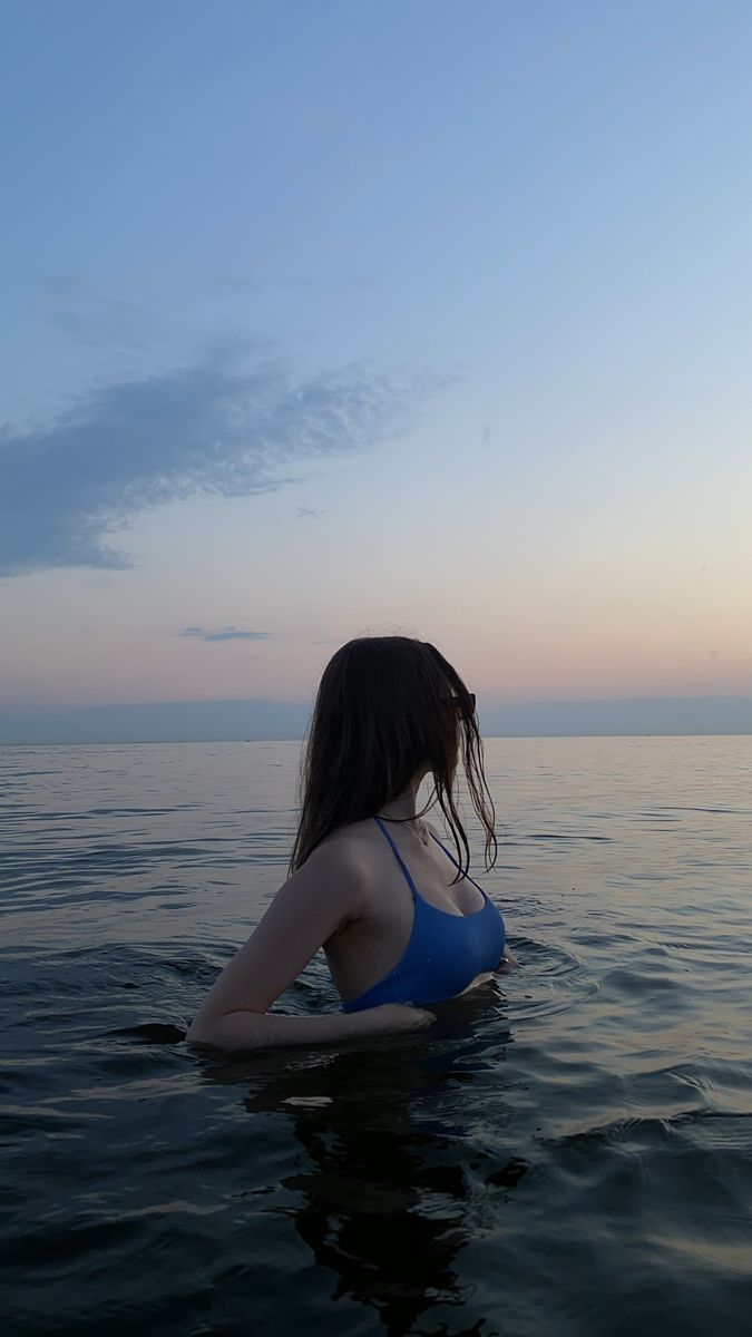 a woman is sitting in the water looking out at the ocean while wearing a blue bathing suit