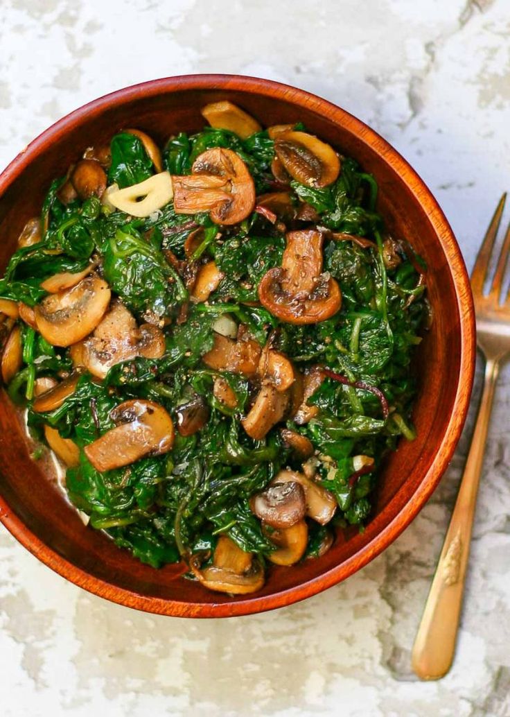 a brown bowl filled with spinach and mushrooms next to a gold fork on top of a table