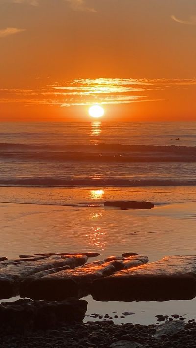 the sun is setting over the ocean with rocks in the foreground and water on the ground