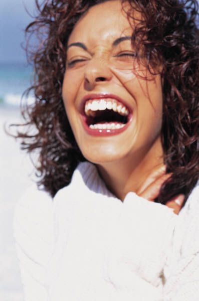 a woman laughing on the beach with her hair blowing in the wind and wearing a white sweater