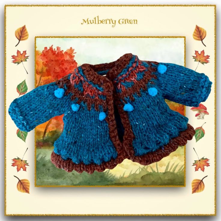 a blue and brown knitted coat with pom poms on it, sitting in front of an autumn scene