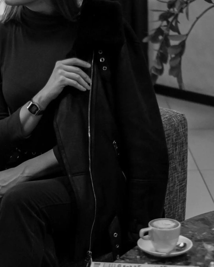 a woman sitting on a couch with a jacket over her shoulders and a cup of coffee in front of her