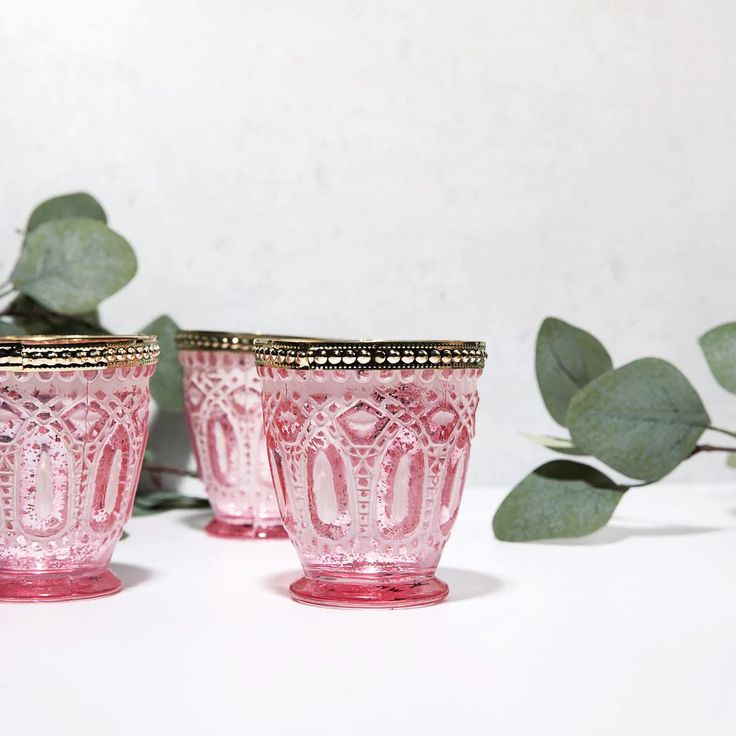 three pink glass cups sitting next to each other on a white surface with green leaves in the background