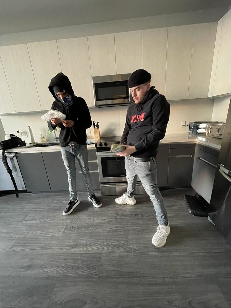 two young men standing in a kitchen next to an oven and microwave, one wearing a black hoodie