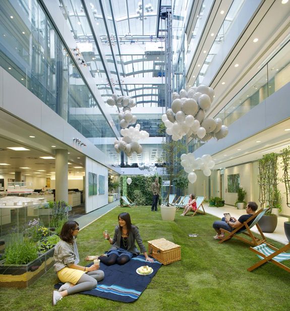 two women sitting on the grass in an office building with balloons hanging from the ceiling