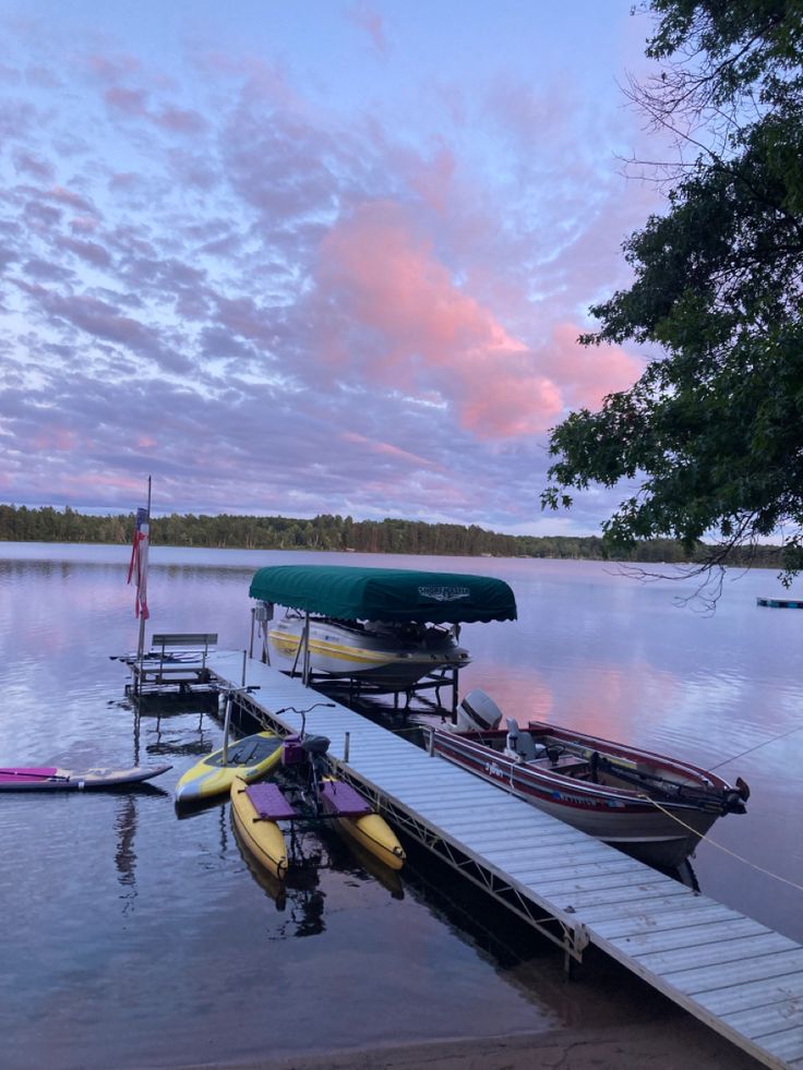 several boats are docked at the end of a pier on a lake with pink clouds in the sky