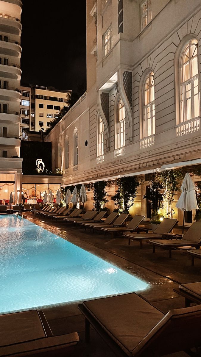 an outdoor swimming pool with lounge chairs and umbrellas around it in front of tall buildings at night