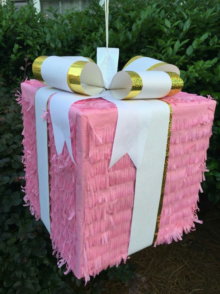 a pink and white gift wrapped in tissue paper with gold foil on top, sitting next to shrubbery