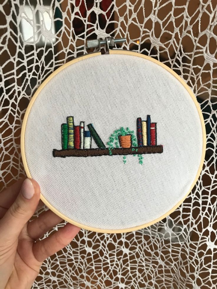 a hand embroidered book shelf with books and plants on it, in front of a lace doily