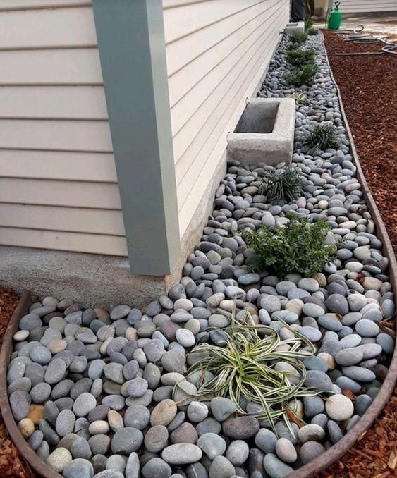 some rocks and plants are in the middle of a garden bed near a house with white siding