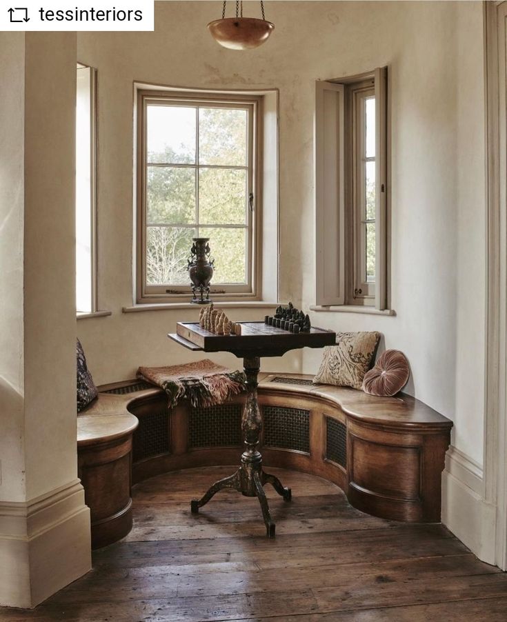 a corner table in the middle of a room with two windows and a bench next to it