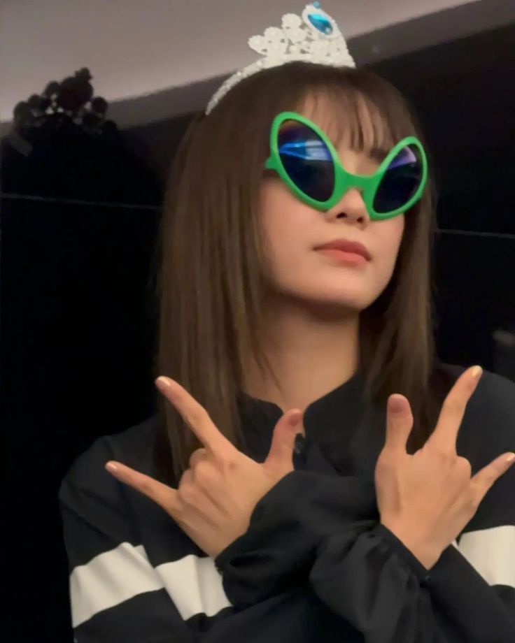 a woman wearing green sunglasses and a tiara making the peace sign with her hands