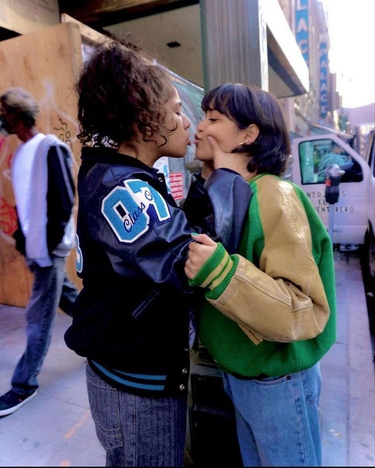 two young women standing next to each other on a sidewalk kissing and smiling at each other