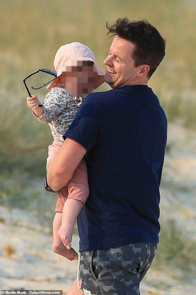 a man holding a baby on the beach with his face covered by sunglasses and smiling